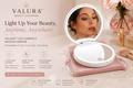 An open Valura LED compact makeup mirror with a built-in light, shown on a pink zip case with a woman's face visible in the circular mirror. The ad includes marketing text, icons, and cosmetic props on a soft beige background.