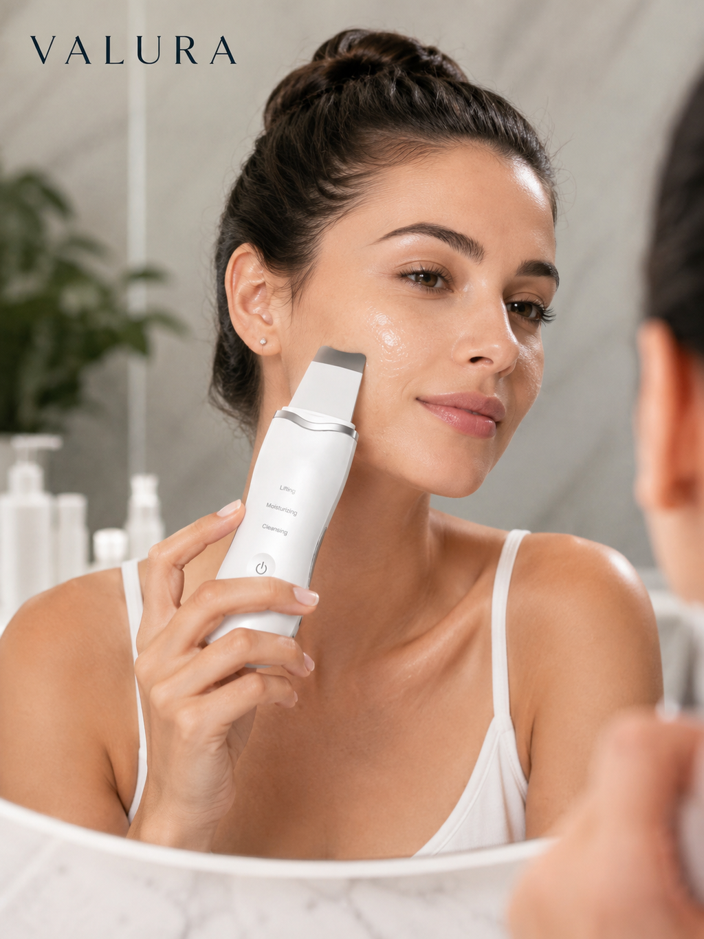 A woman in a white tank top holds a white Valura facial skin treatment device against her cheek in a bright bathroom setting, with her reflection visible in the mirror.