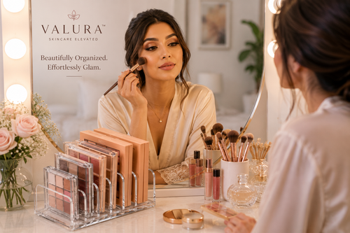 A woman in a beige robe sits at a vanity mirror applying makeup, with Valura skincare products displayed on the counter and the Valura brand logo visible on the wall. The scene includes makeup palettes in a clear stand, brushes in a cup, and soft pink flowers on the left.